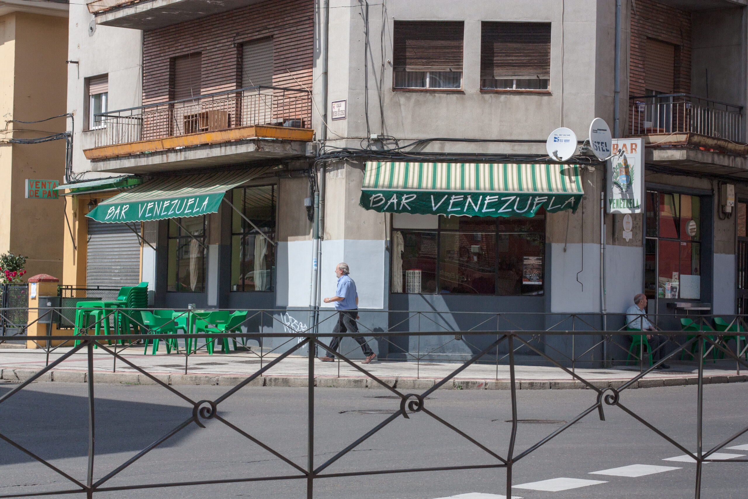 En la Esquina de al frente, diagonal, un bar con el nombre de Bar Venezuela, un señor caminando a la izquierda, otro sentado en la sombra de lado derecho de la imagen, color del nombre y y las sillas del lugar son del color verde.